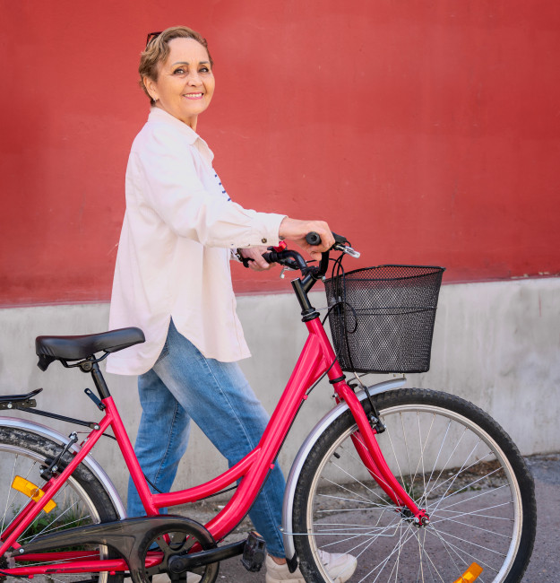 Foto: ältere, weiße Frau mit kurzen Haaren schiebt ein Fahrrad vor einer roten Hauswand und lächelt